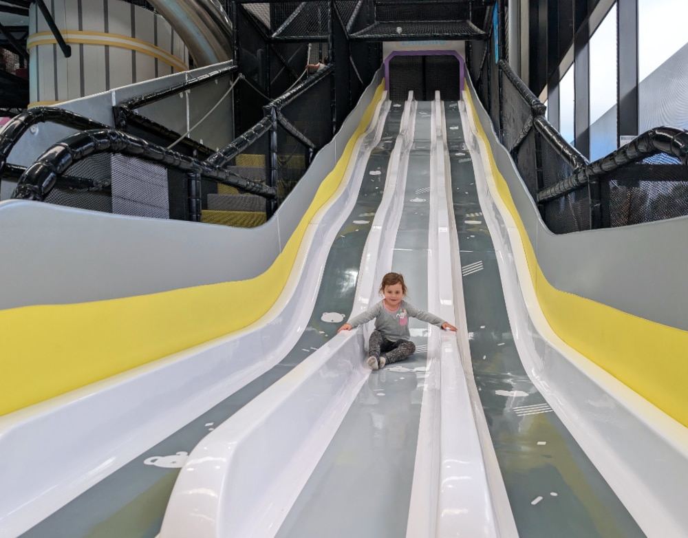 NEW Landing Play Centre Altona North 3 Child sitting on a multi-lane indoor slide inside Landing Play Centre Altona North, with high sides and netted play structure around it